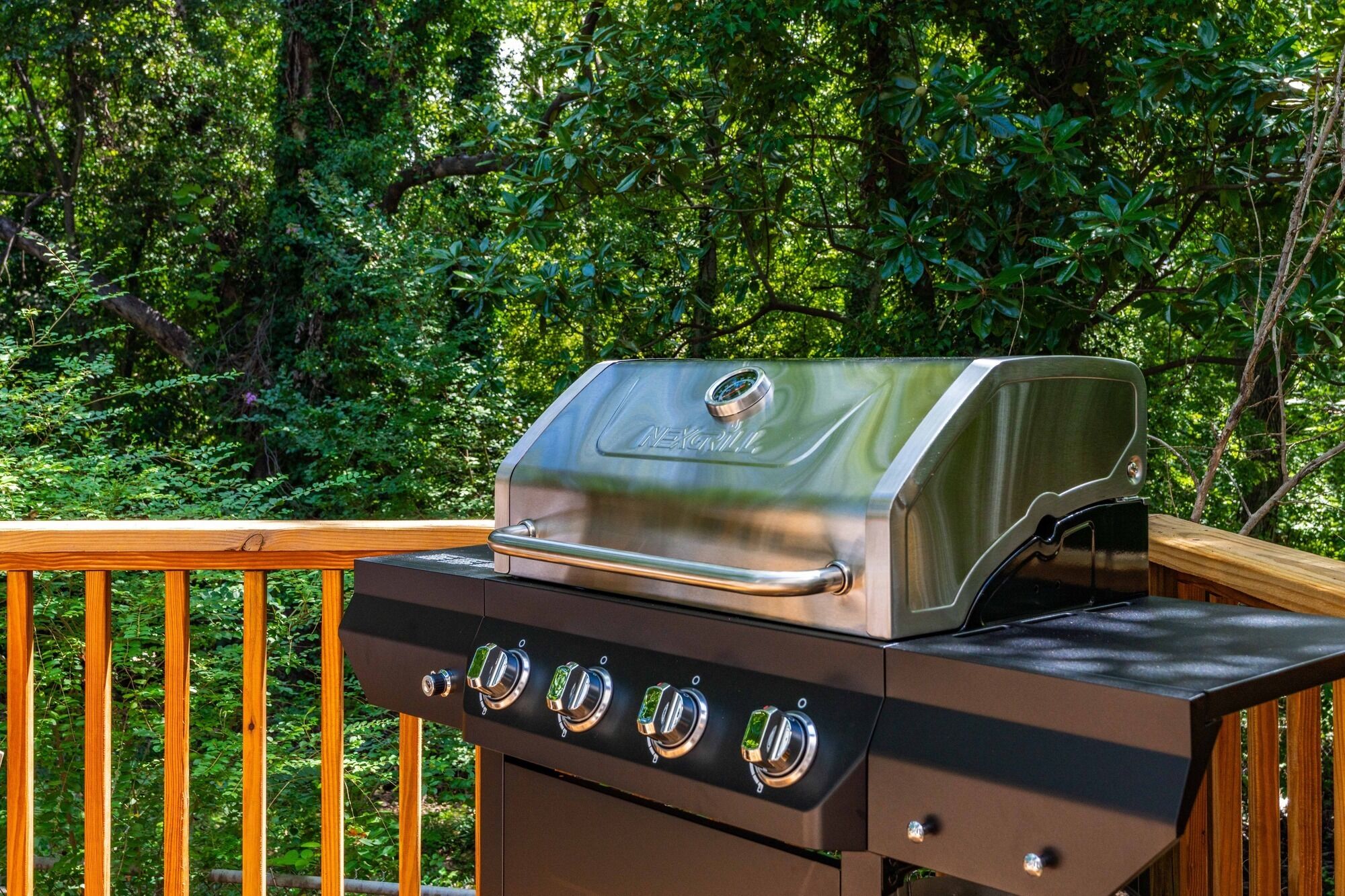 A stainless steel grill with four burners, set on a wooden deck with a forest backdrop.