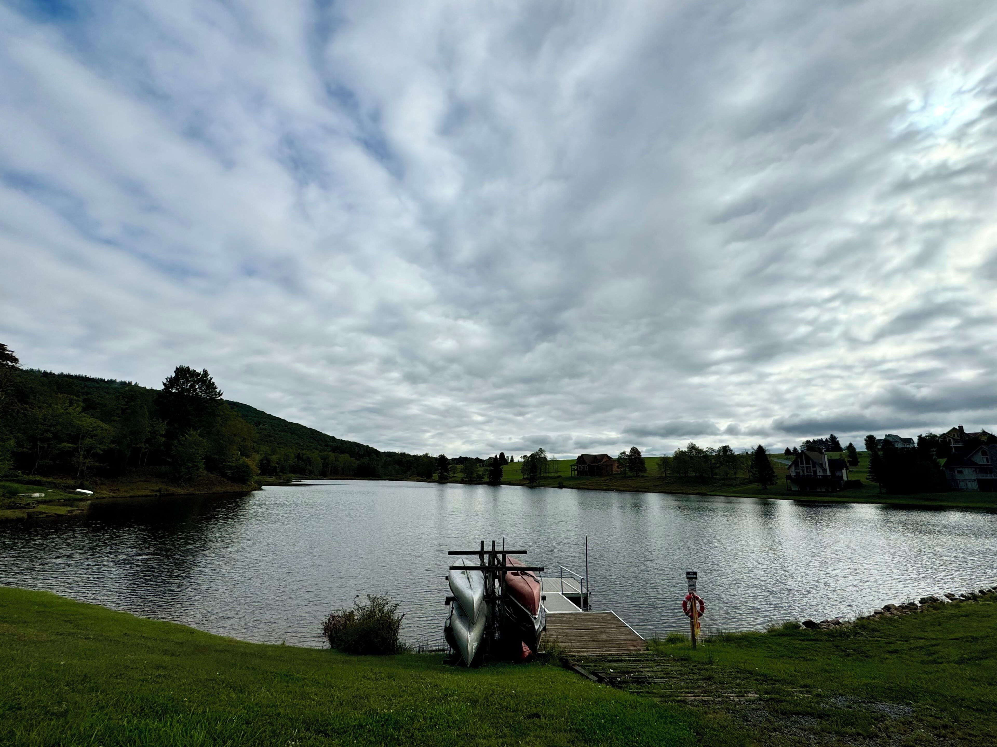 A calm lake with a dock and boats, surrounded by greenery and houses in the distance.