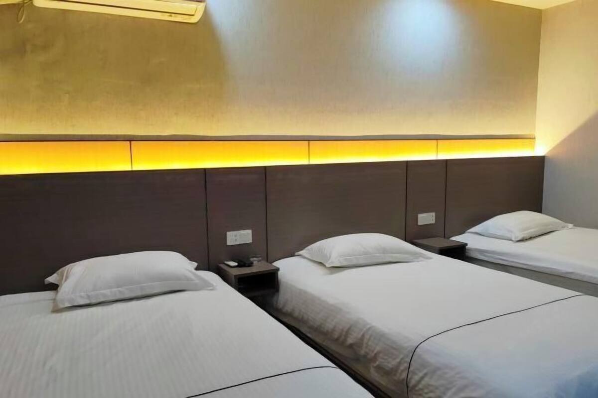 Habitació d'hotel amb tres llits individuals, un capçal de llit amb il·luminació integrada, una petita taula de nit i un aparell d'aire condicionat fixat a la paret.
