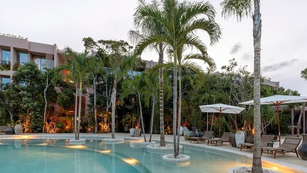 Une piscine entourée de palmiers et d’un bâtiment doté de balcons, avec des chaises longues et des parasols à proximité.