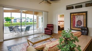 Ein geräumiges Wohnzimmer mit Glastür zum Balkon, Deckenventilator, rotem Ledersessel, Glastisch und einem gerahmten Kunstwerk an der Wand.
