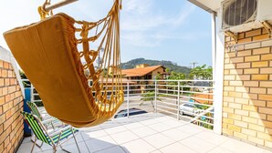 Un balcón con una silla colgante, una silla de metal y vistas a una zona residencial con casas y un paisaje montañoso.