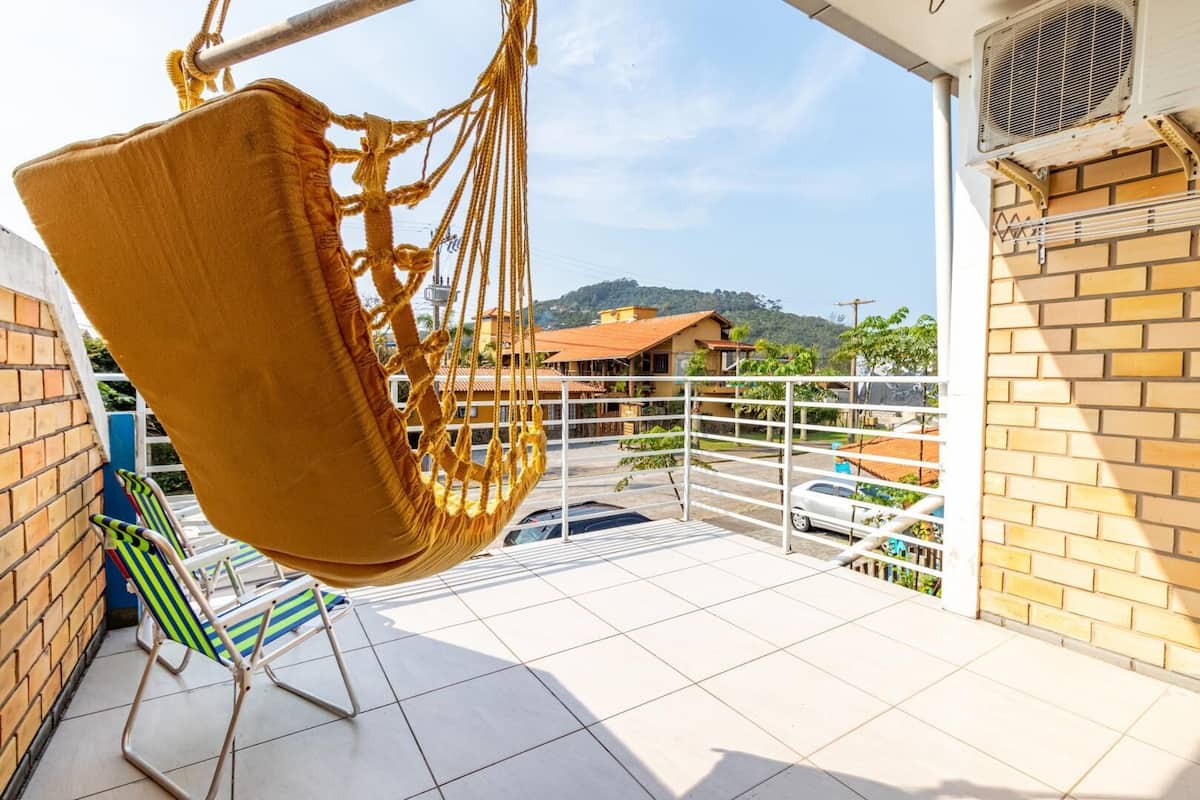 A balcony with a hanging chair, a metal chair, and a view of a residential area with houses and a hilly landscape.
