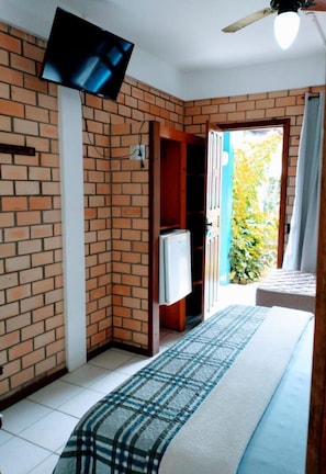 A hotel room with a brick wall, a bed with a plaid bedspread, a television mounted on the wall, and a ceiling fan. - Pousada Baleia Franca (Garopaba)