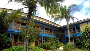 Un edificio de dos plantas con paredes azules y color beige, rodeado de abundante follaje y palmeras.