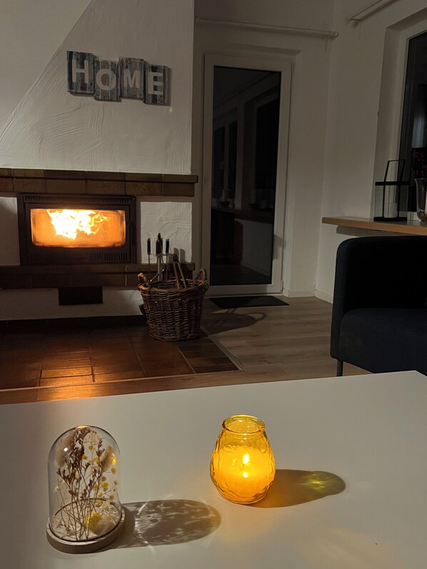 A living room with a fireplace, a 'HOME' sign, a wicker basket, a glass dome with dried flowers, and a lit candle. - Ferienhaus Seeblick am Bitburger See! (Biersdorf am See)
