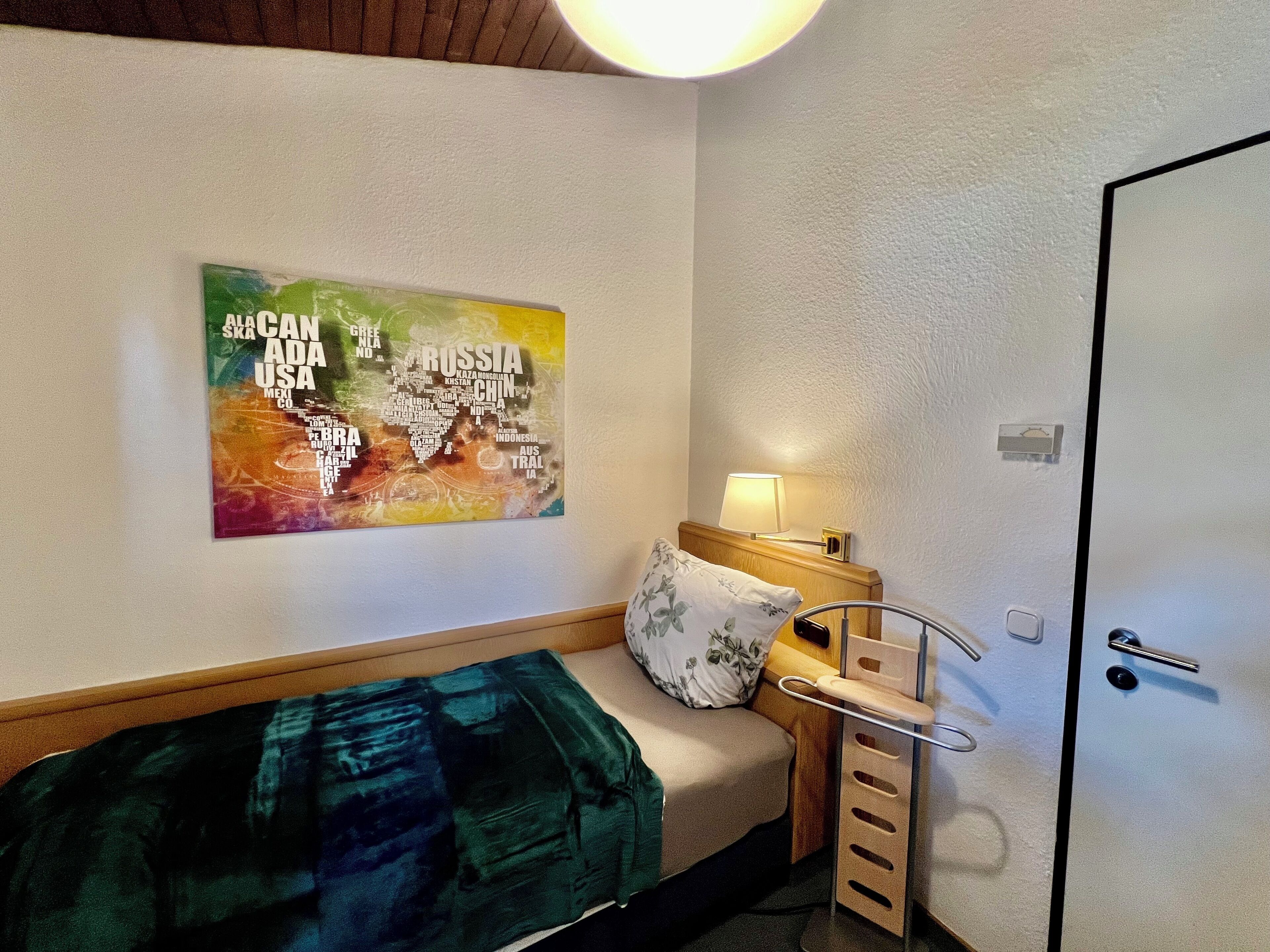 Et lille, pænt indrettet rum med en enkelt seng, et natbord med lampe og et kort ophængt på væggen.