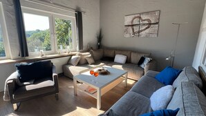 En stue med sofa, lænestol og sofabord. Der er et vindue med gardiner, et billede på væggen og en plante.