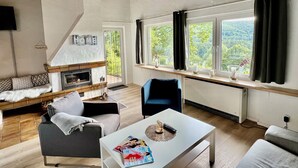 En moderne stue med pejs, sofa, sofabord og udsigt til naturen gennem store vinduer.
