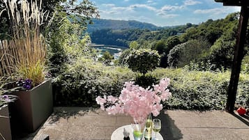 A patio with wicker furniture, a table with a vase of flowers, and a view of a lake and hills.