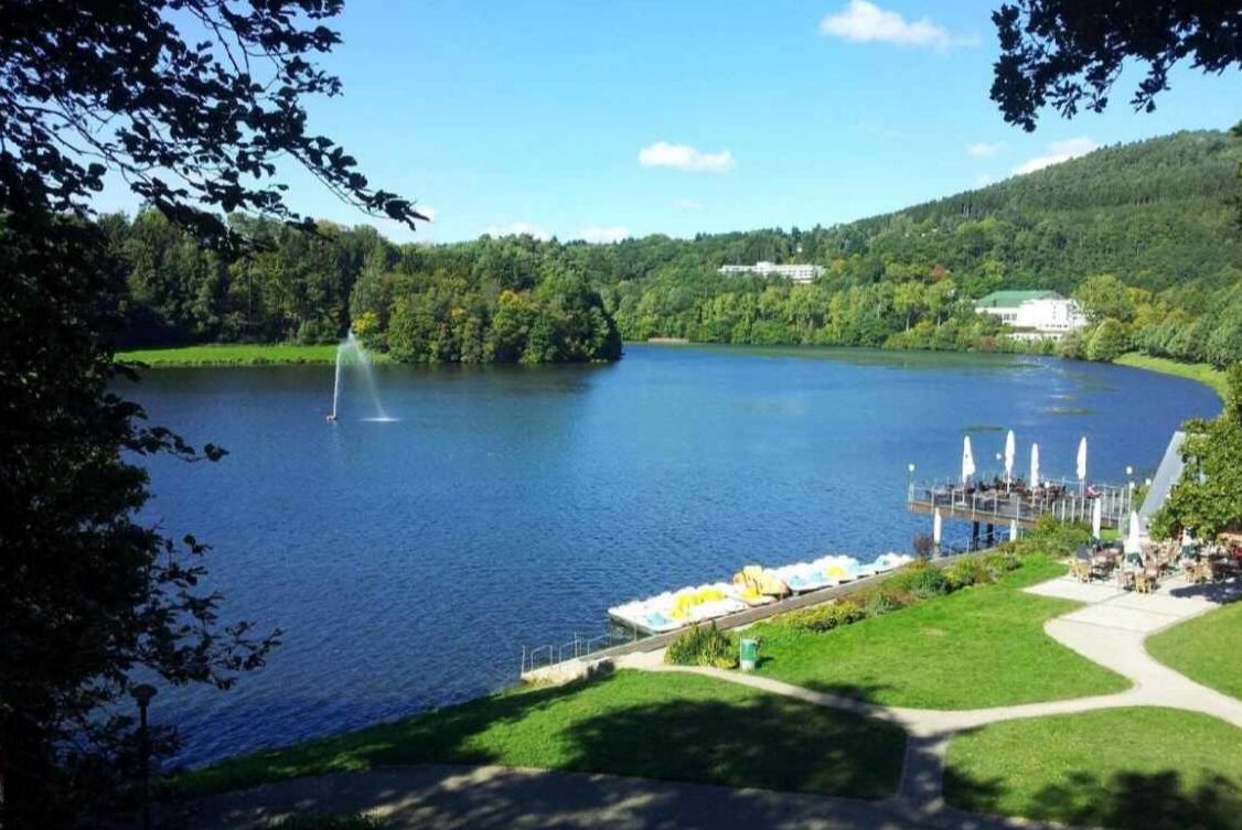 A lake with a fountain, surrounded by greenery and a dock with umbrellas.