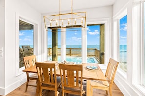 A dining area with a wooden table and chairs, a hanging light fixture, and a view of a pool and beach through large windows. - Beachfront | Heated Pool | Hot Tub | Fenced Yard with Game Area (Crystal Beach)