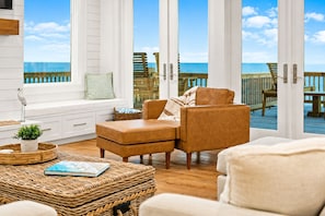 A room with a view of the ocean, featuring a sofa, a wicker ottoman, and a window with a view of a wooden deck. - Beachfront | Heated Pool | Hot Tub | Fenced Yard with Game Area (Crystal Beach)