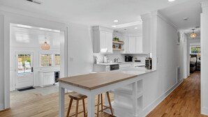 A modern kitchen with white cabinetry, a wooden island with stools, and a view into the adjacent living area. - 16 29th Avenue Isle of Palms Group Retreat (Isle of Palms)
