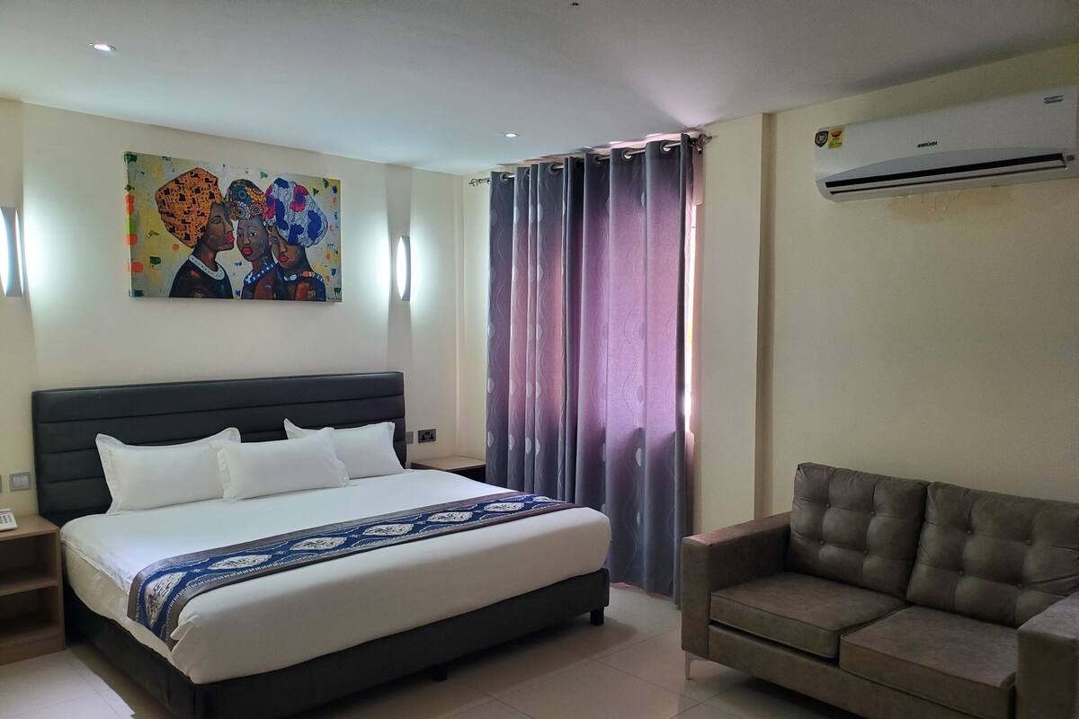 A hotel room with a large bed, a sofa, a painting on the wall, and an air conditioning unit.