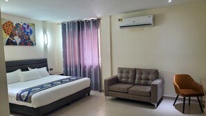A hotel room with a bed, a sofa, a chair, and an air conditioning unit.