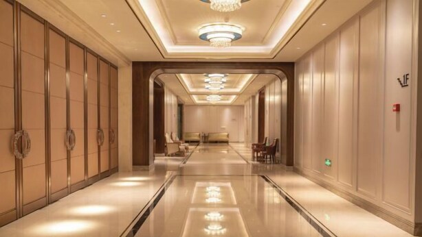 Un couloir d’hôtel avec un sol réfléchissant, un lustre et un espace détente au bout.