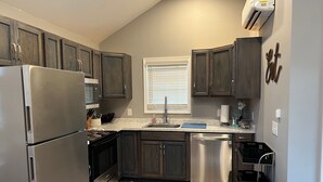 A compact kitchen with dark wooden cabinets, stainless steel appliances, a window with blinds, and a small trash bin.