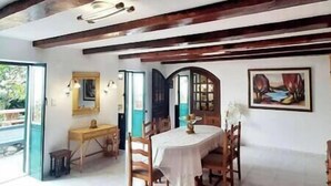 Un coin repas avec une table et des chaises en bois, un tableau accroché au mur et une vue sur l’extérieur par une porte ouverte.