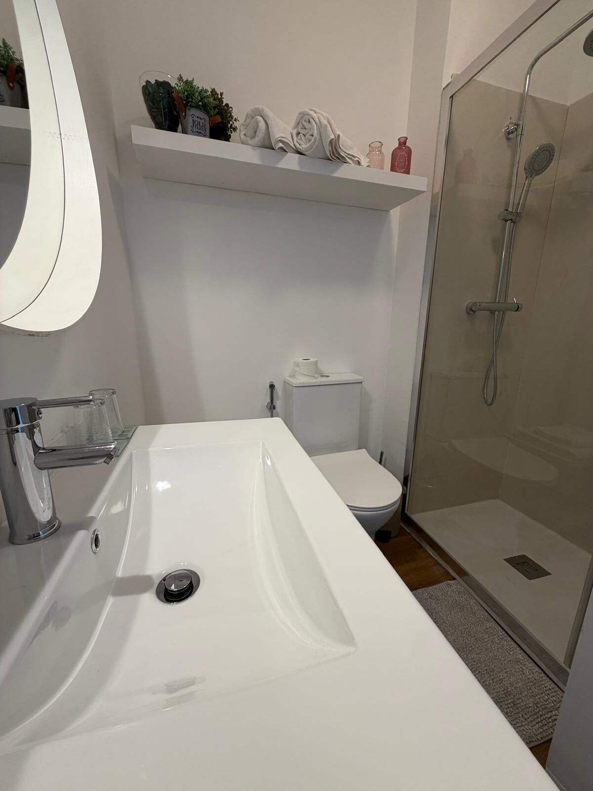 A bathroom with a white sink, toilet, and shower. There is a shelf above the toilet with decorative items.