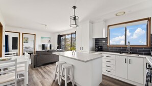 Een moderne keuken met witte kasten, een groot eiland met krukken, een eethoek met een witte tafel en stoelen, en een uitzicht op het buitengebied door een groot raam.
