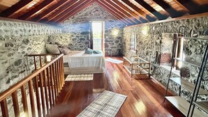 Quarto de sótão com teto de madeira, paredes de pedra, uma cama e uma porta de vidro que dá para o exterior.