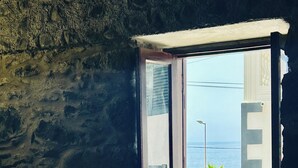 Quarto com parede de pedra, janela com vista para o mar e cama com cobertor branco.