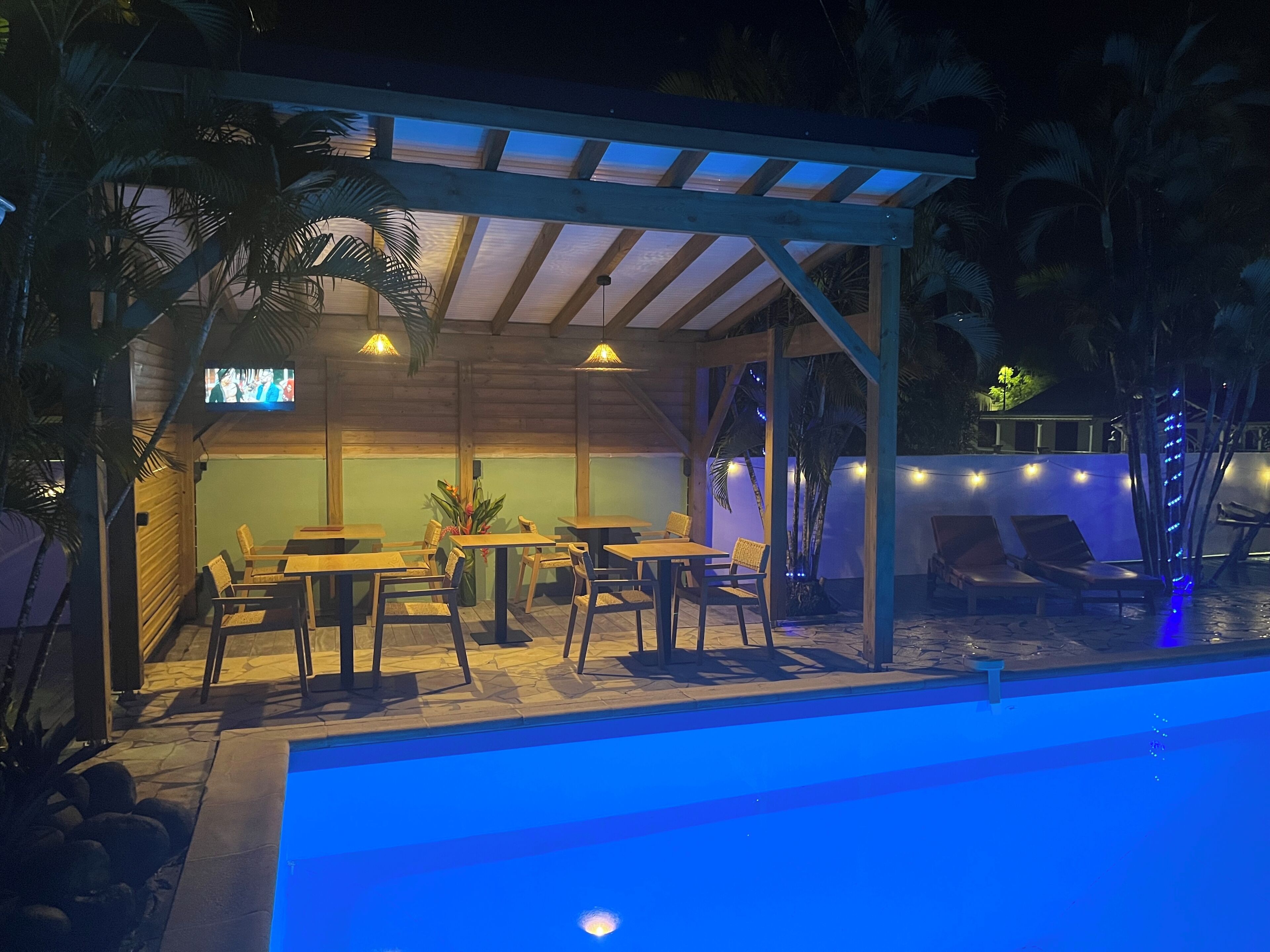 An outdoor pool area with wooden tables and chairs, a covered seating area, and a TV screen.