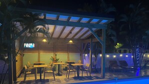 Ein Poolbereich im Freien mit Holztischen und -stühlen, einer überdachten Sitzgelegenheit und einem Fernsehbildschirm.