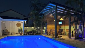 Ein Schwimmbad mit Holzterrasse, Sitzgelegenheiten im Freien und einem Fernsehbildschirm.