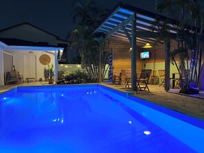 A swimming pool with a wooden deck, outdoor seating, and a TV screen. - Chambre Studio Confort, Plein Centre, Accès Piscine, Idéal Pour Couple (Baie-Mahault)