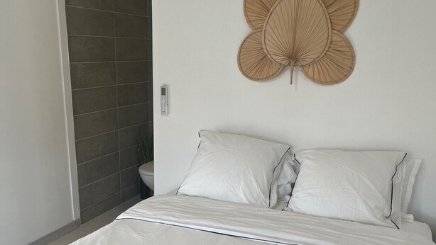 Ein ordentlich bezogenes Bett mit weißen Leinen und zwei zusammengerollten Handtüchern, ein dekorativer Wandventilator und eine Tür zu einem Badezimmer.