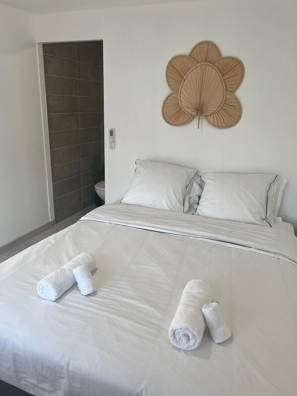 A neatly made bed with white linens and two rolled towels, a wall-mounted decorative fan, and a door leading to a bathroom. - Chambre Studio Confort, Plein Centre, Accès Piscine, Idéal Pour Couple (Baie-Mahault)