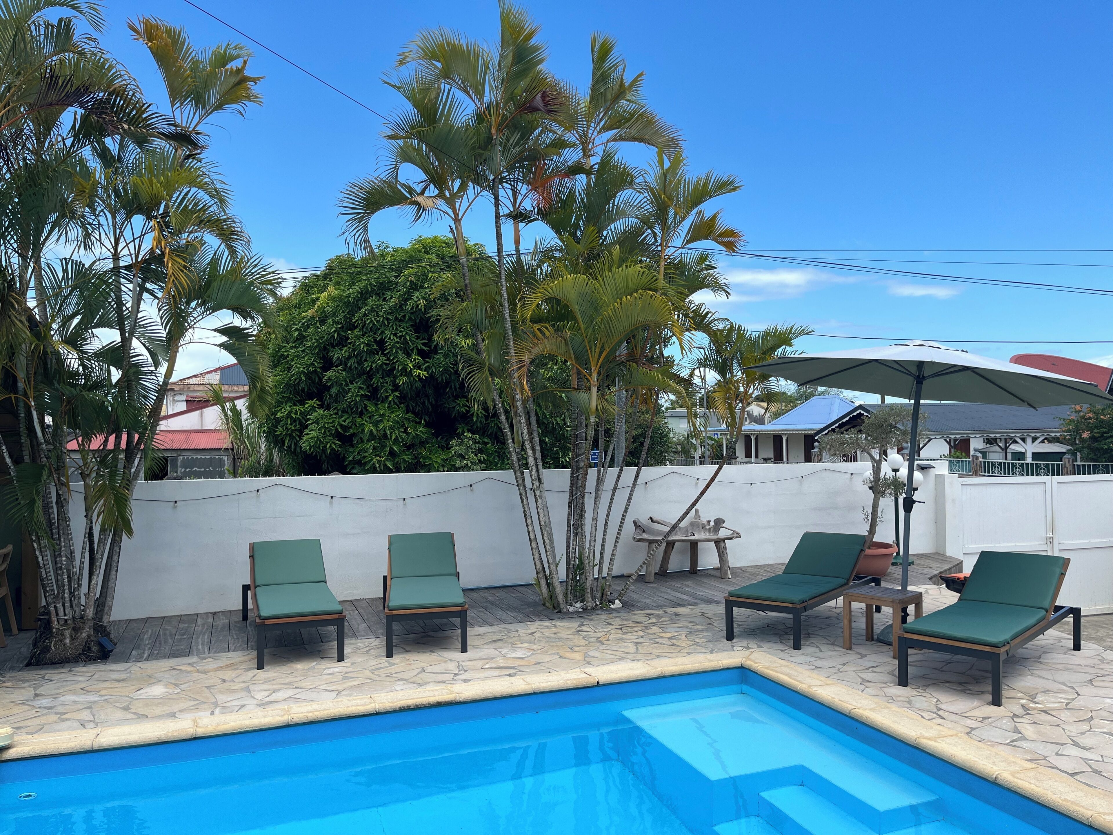 A swimming pool with lounge chairs and a table, surrounded by palm trees and a white wall.