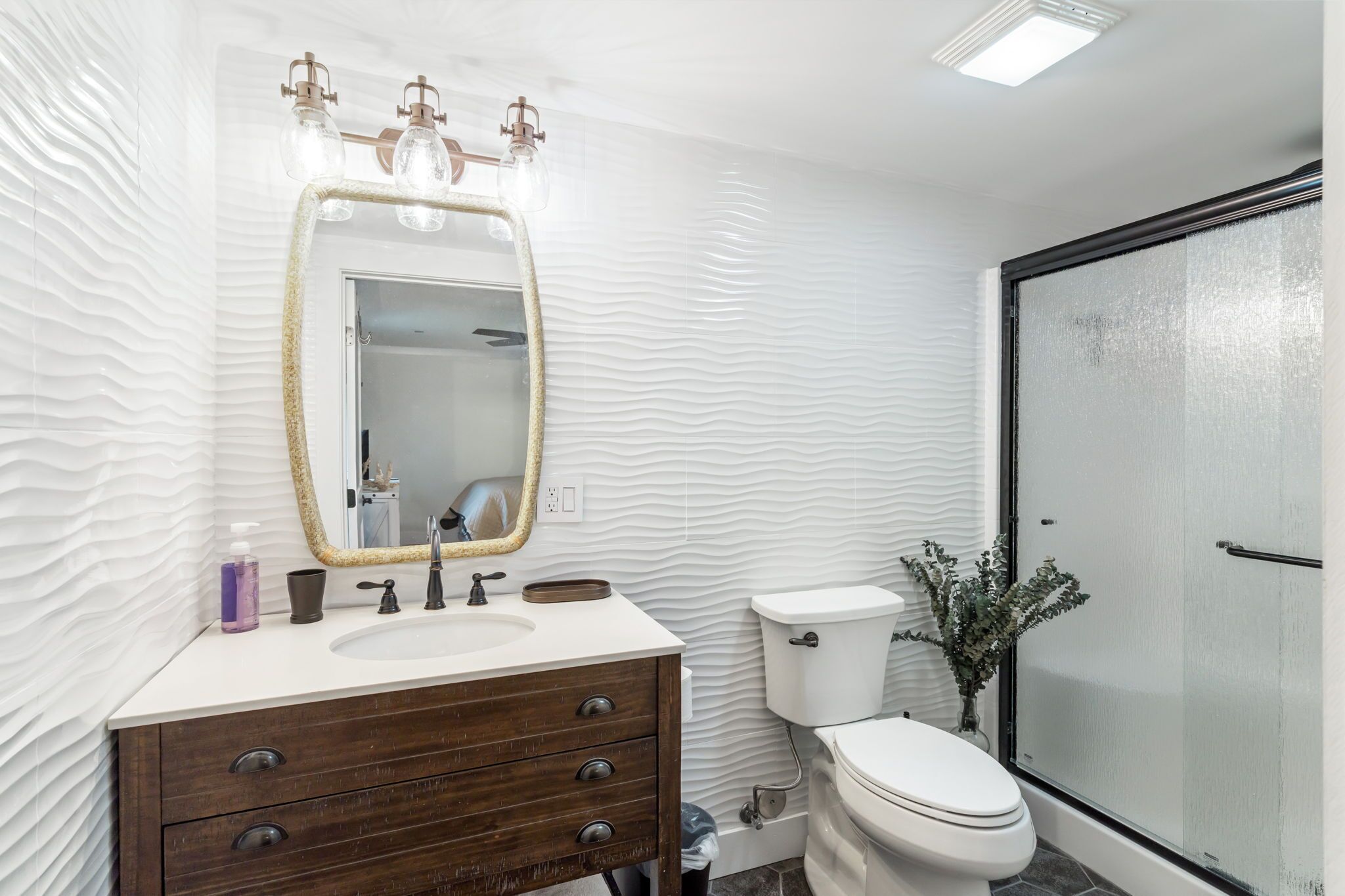 Un baño con pared blanca texturizada, un tocador de madera con espejo, inodoro y zona de ducha.