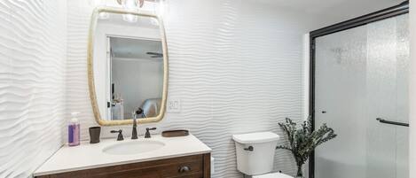 Un baño con pared blanca texturizada, un tocador de madera con espejo, inodoro y zona de ducha.