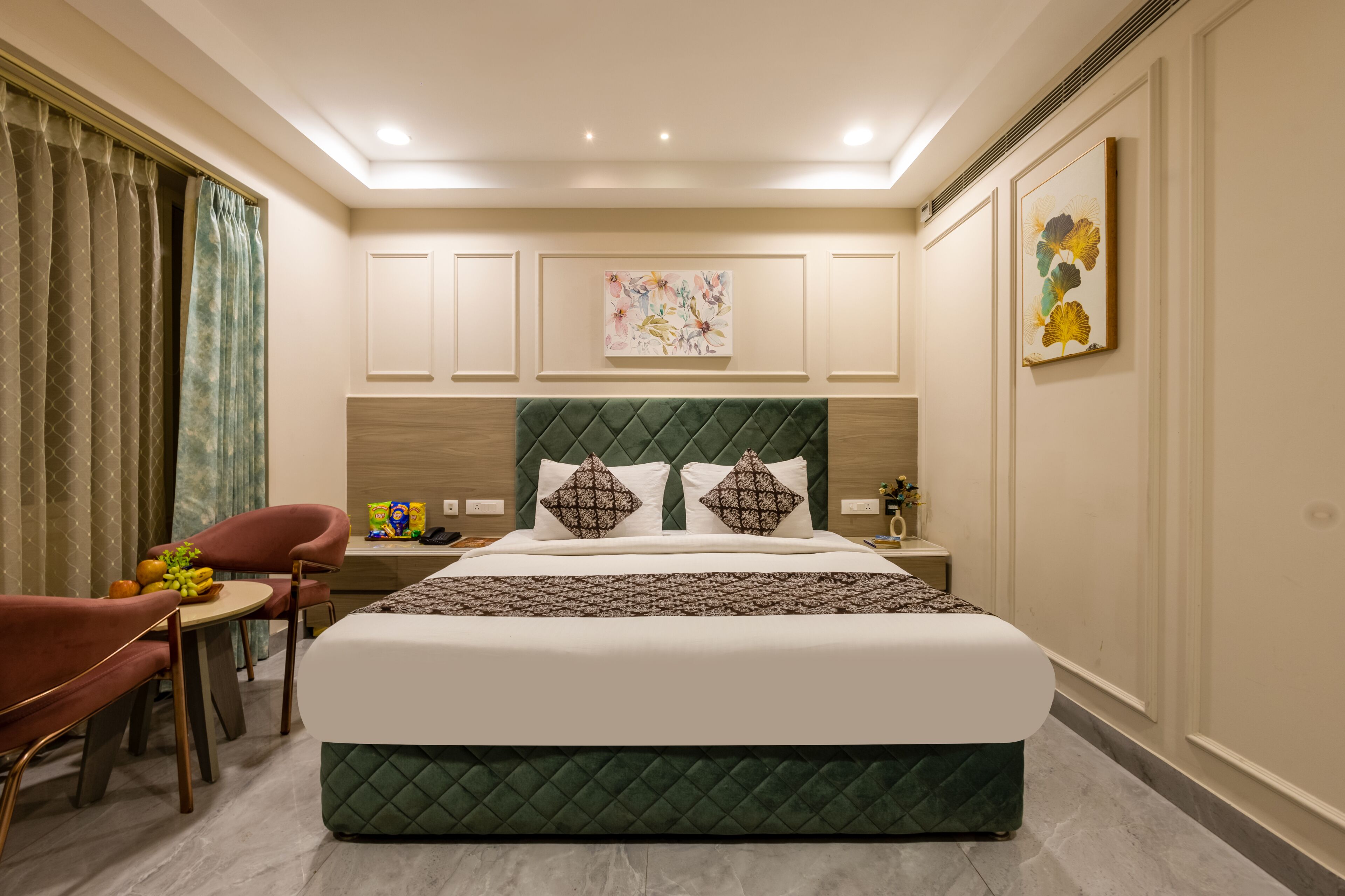 Une chambre d’hôtel avec un lit, une chaise, une table avec des fruits, une table de chevet et des œuvres d’art accrochées aux murs.