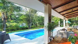 A modern outdoor pool area with a wooden deck, a seating area with a sofa and chairs, and a view of lush greenery.