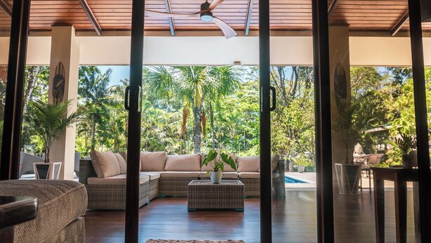 A modern living room with a large glass door leading to a pool area, surrounded by lush greenery.
