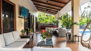 A spacious living area with a wooden ceiling, comfortable seating, a flat-screen TV, and a view of a swimming pool.