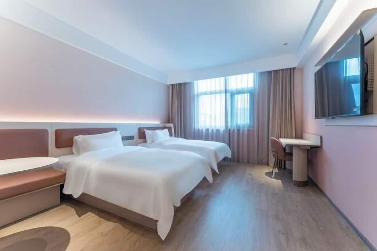 A hotel room with two beds, a desk, a chair, a TV, and large windows with curtains.
