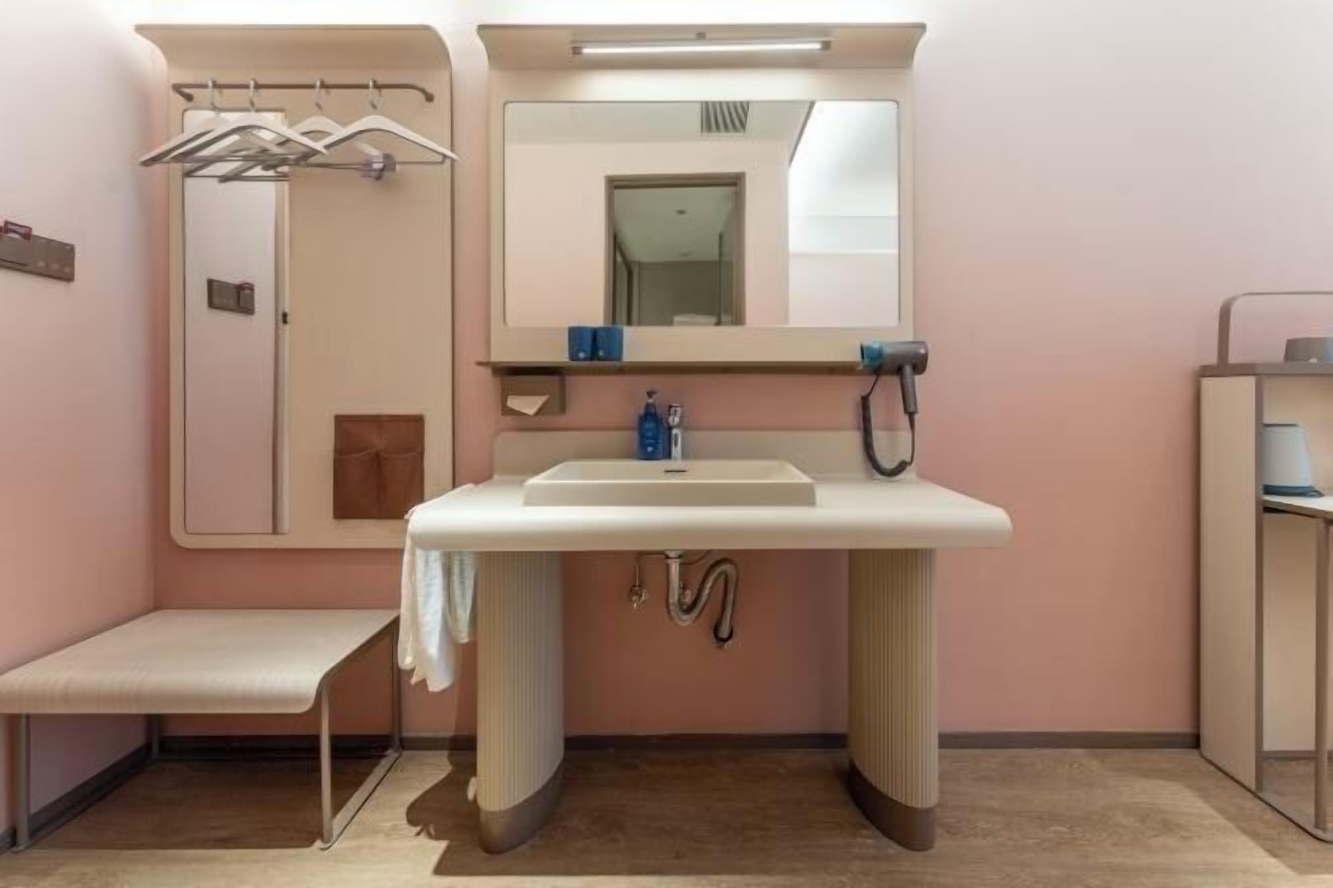 Un baño con lavamanos, espejo, toallero, banco y una puerta que da a otra habitación.