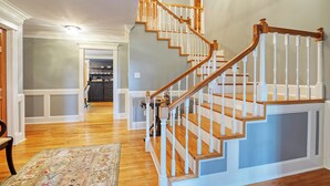 Un escalier avec des marches en bois et des balustrades blanches, menant à un palier d’où l’on peut apercevoir une cuisine et une salle à manger.