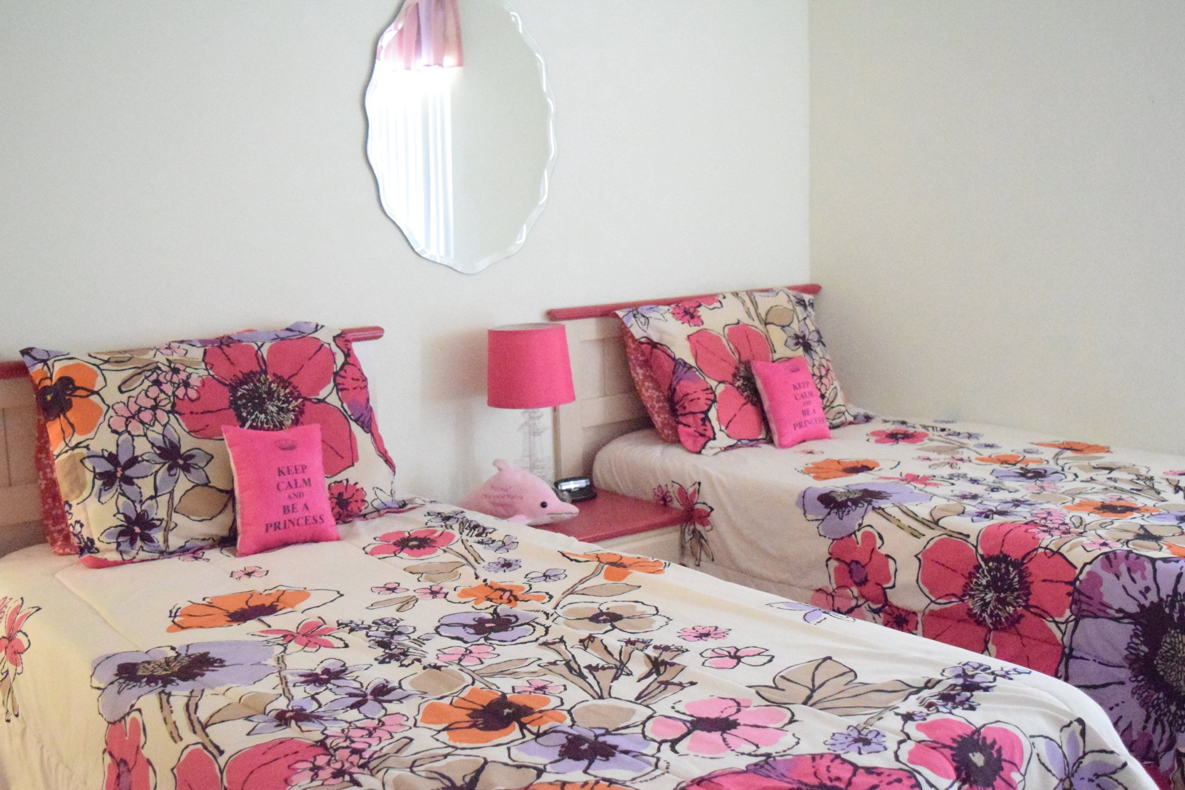 A bedroom with two beds, floral bedding, pink pillows, a mirror, and a pink lamp.