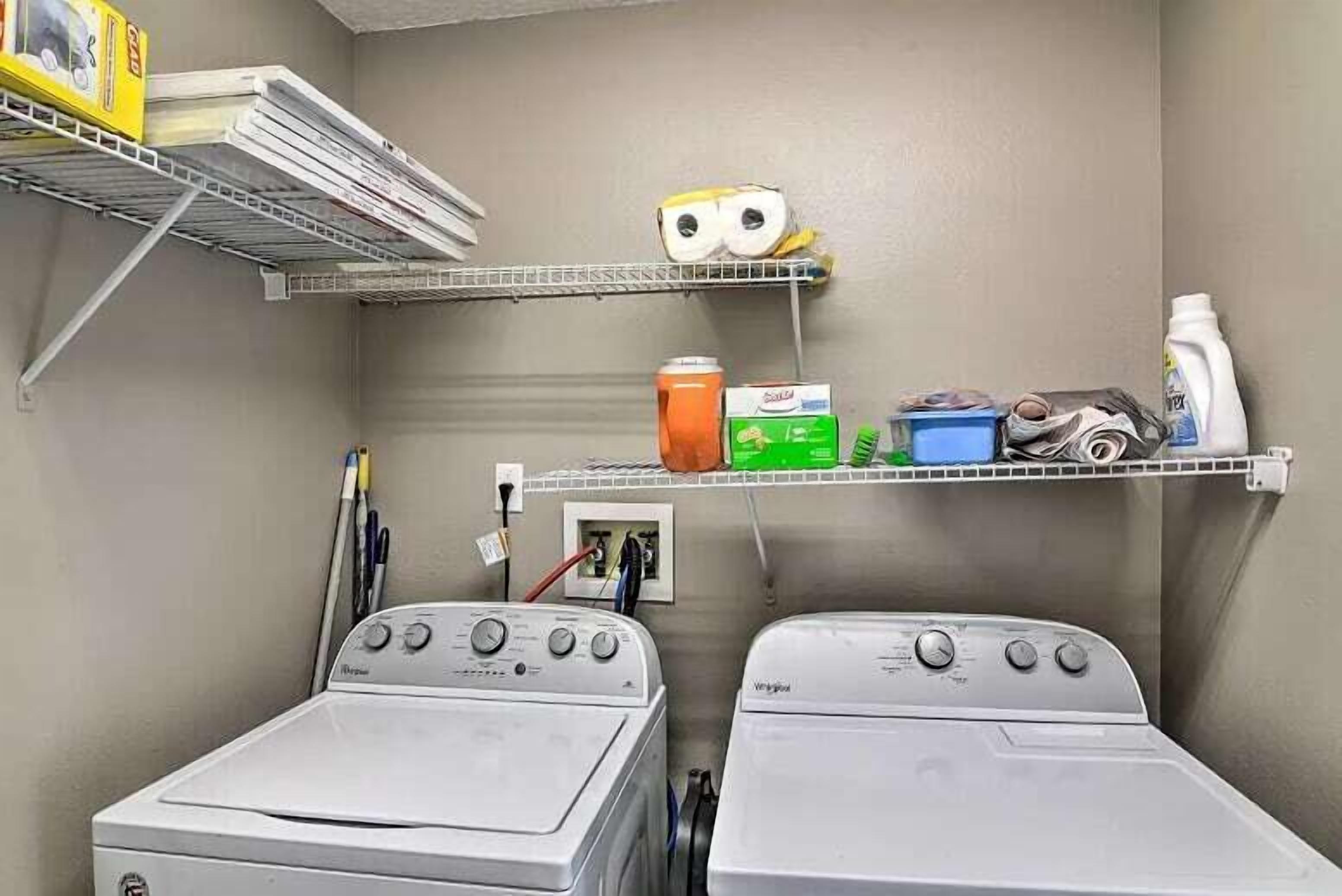 A laundry room with two front-loading washing machines, shelves with cleaning supplies, and a wall-mounted shelf with a rolled-up towel and a can of soda.