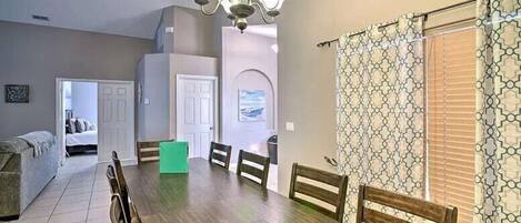 A dining area with a wooden table, chairs, and a chandelier. There is a couch and a door leading to another room.