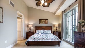 Une chambre avec un grand lit, deux tables de chevet avec des lampes, un ventilateur de plafond, une grande fenêtre avec des rideaux et un tableau encadré au mur.