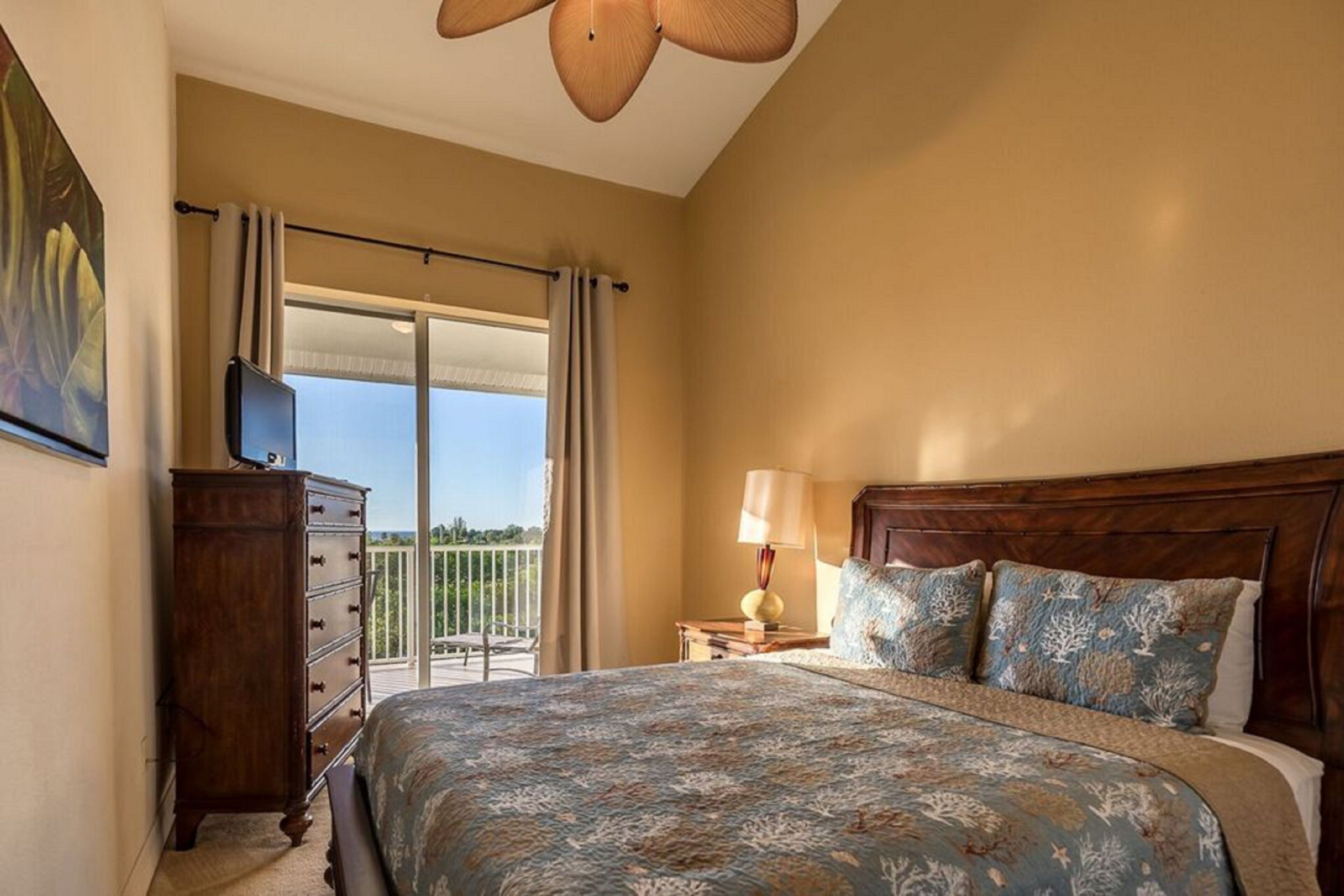 A bedroom with a large bed, a dresser, a nightstand with a lamp, and a view of the outdoors.