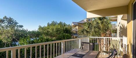 A balcony with a table and chairs, surrounded by a white railing, overlooking a residential area with trees and a building in the background.