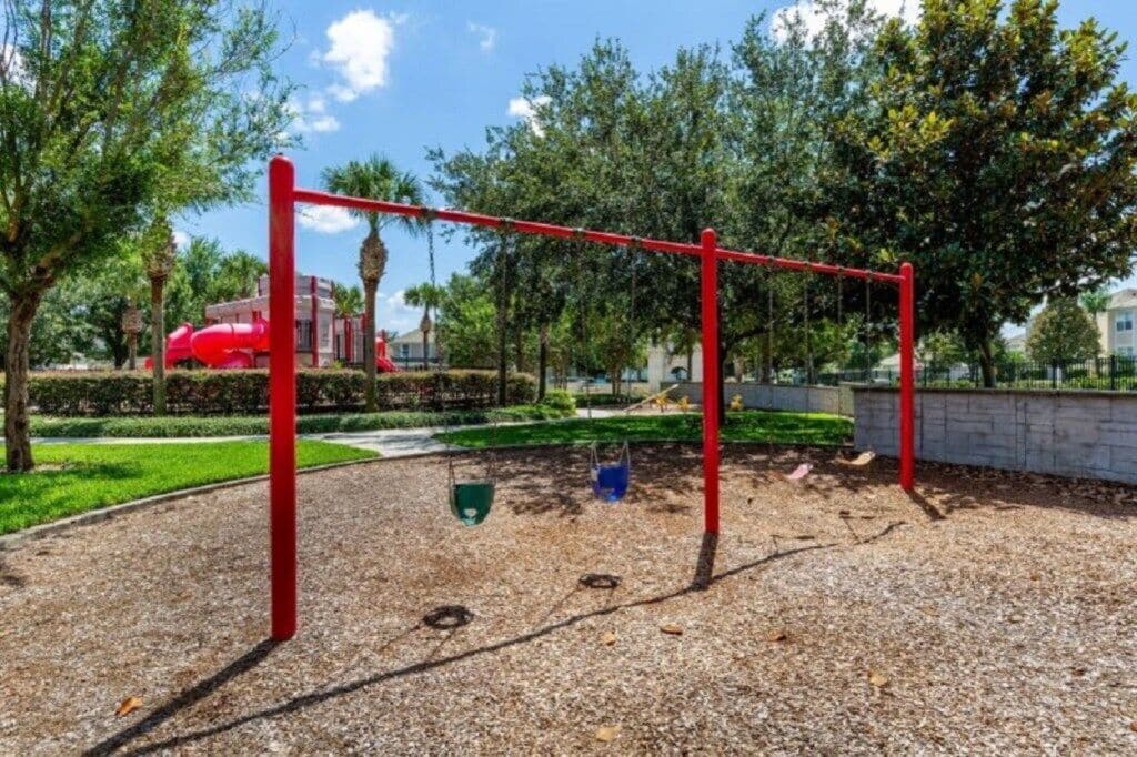 Un parque infantil con un tobogán y columpio rojos, rodeado de árboles y un edificio al fondo.